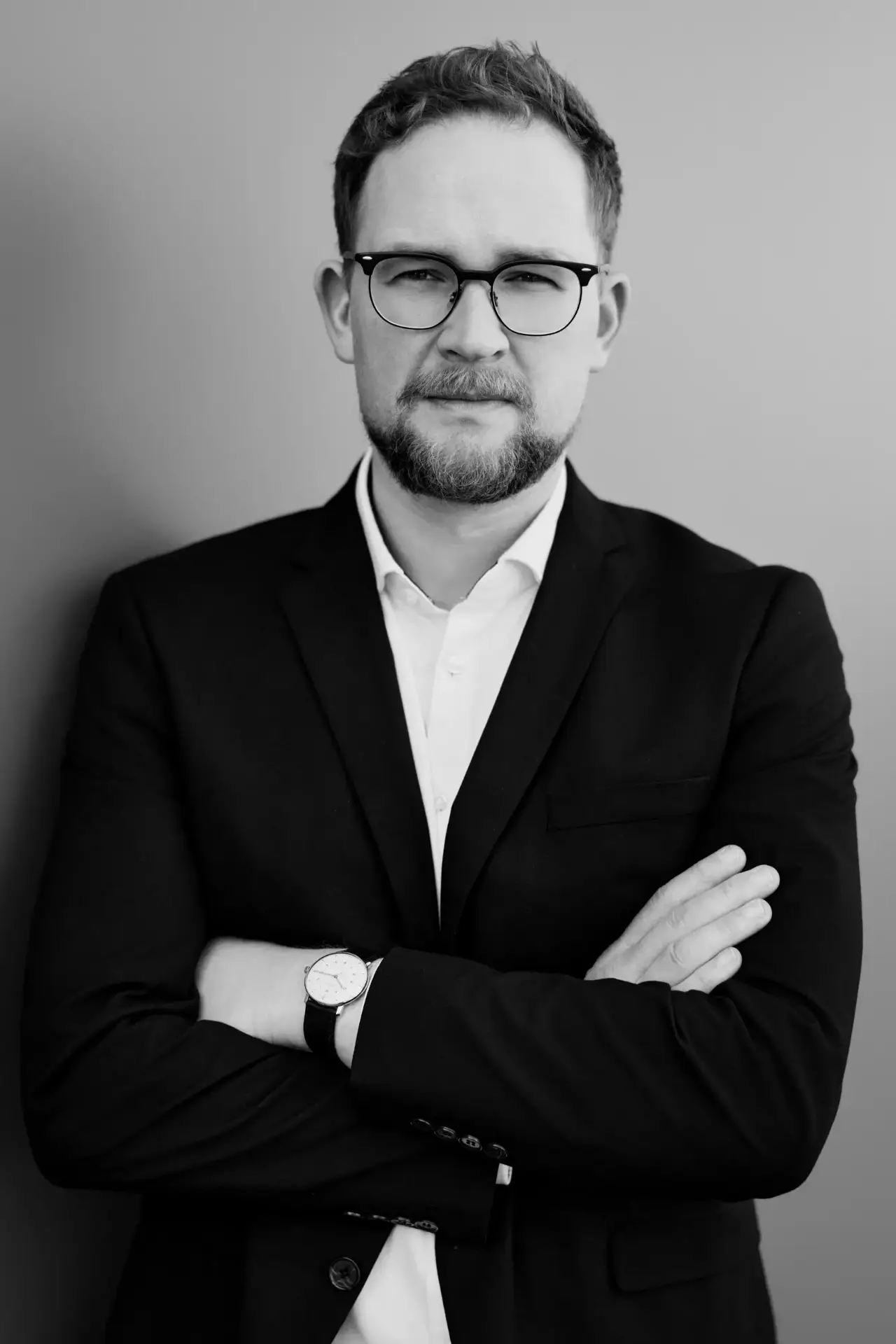 Schwarzweiß-Businessportrait eines Mannes mit Brille, Bart und dunklem Sakko mit verschränkten Armen vor grauer Wand in Hamburg