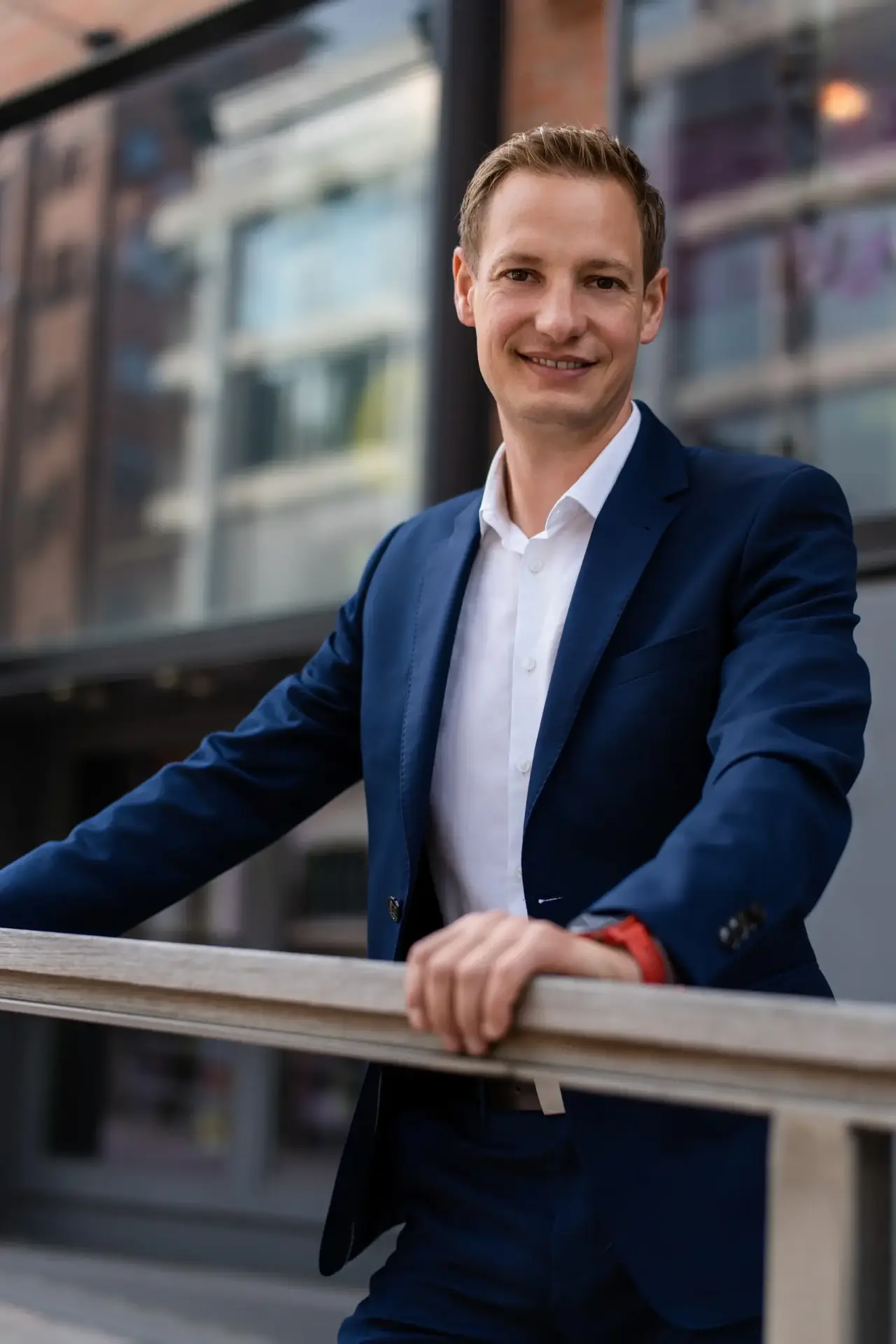 Businessportrait eines lächelnden Mannes im blauen Anzug an einem Geländer vor moderner Architektur in Hamburg