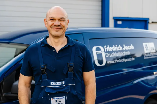 Mitarbeiterfoto Monteur in Arbeitskleidung vor blauem Transporter